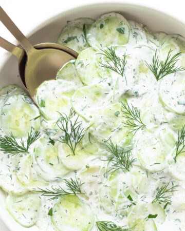 Close up of bowl containing creamy German cucumber salad (Gurkensalat) with gold salad servers.