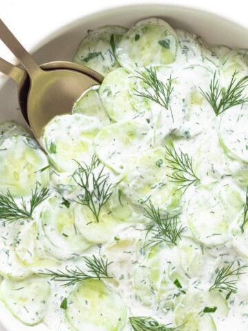 Close up of bowl containing creamy German cucumber salad (Gurkensalat) with gold salad servers.