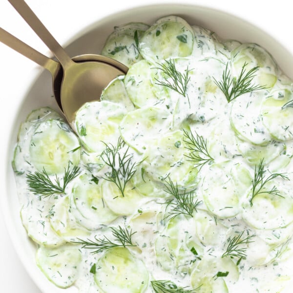 Close up of bowl containing creamy German cucumber salad (Gurkensalat) with gold salad servers.