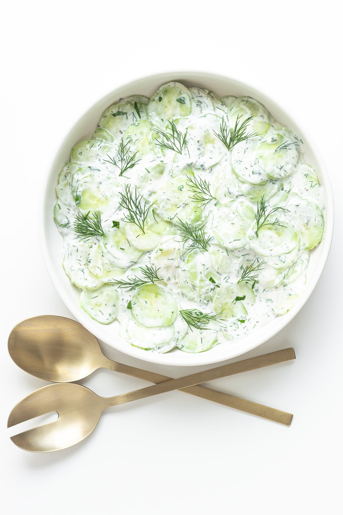 Cucumber salad in a white bowl with gold salad servers next to it.