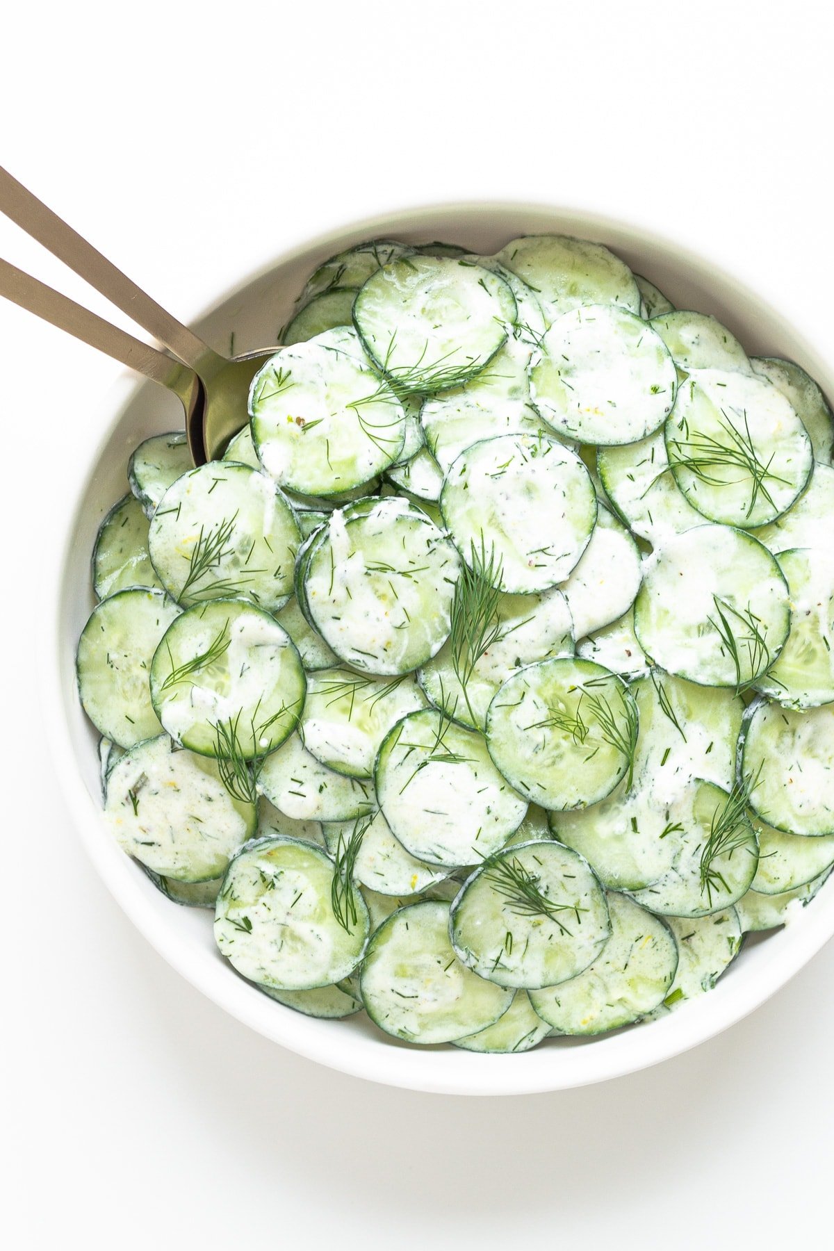 White bowl of greek yogurt cucumber salad with gold serving spoons.