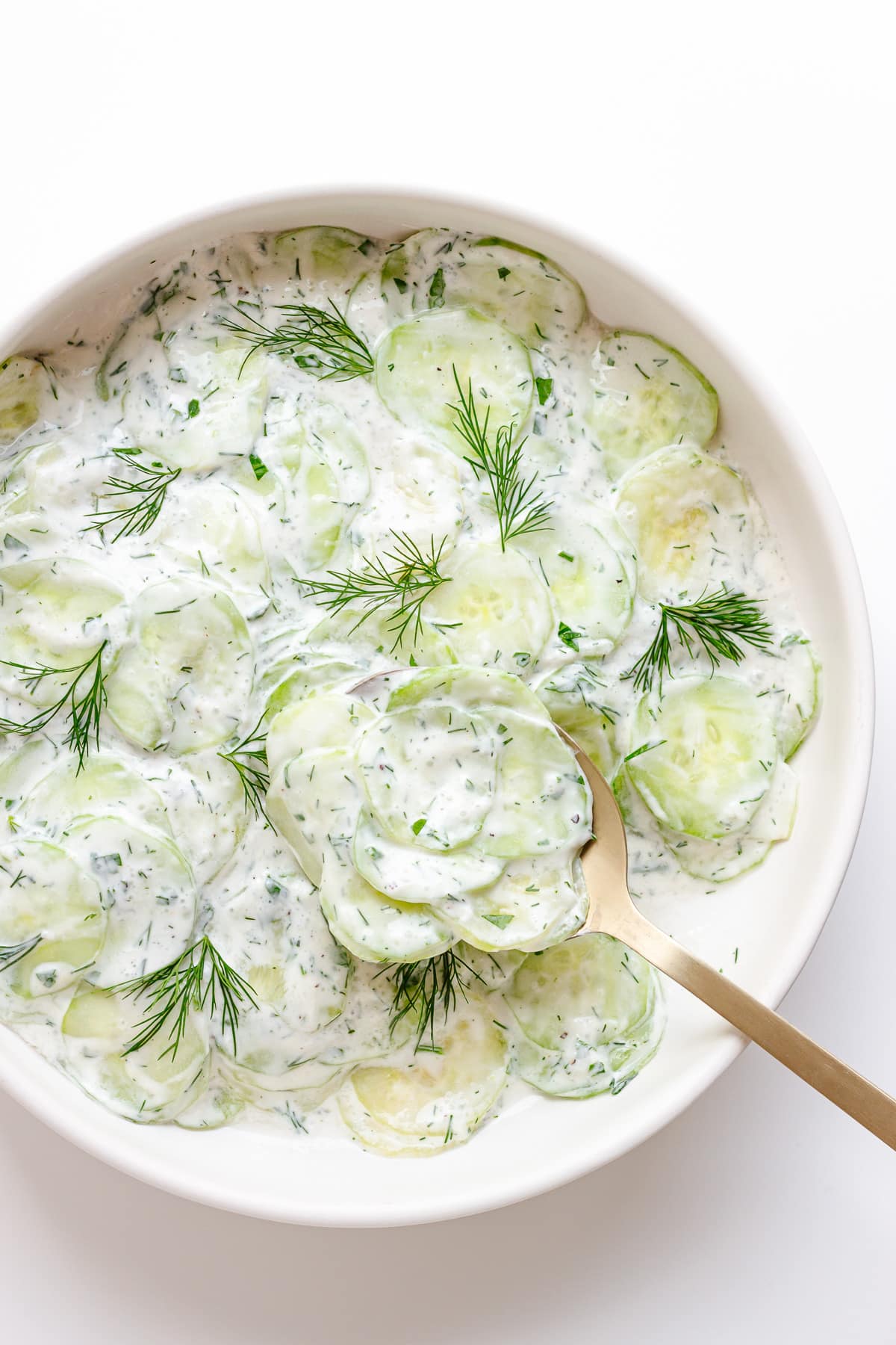 Gurkensalat (creamy German cucumber salad) in a white bowl with gold salad server lifting some out.