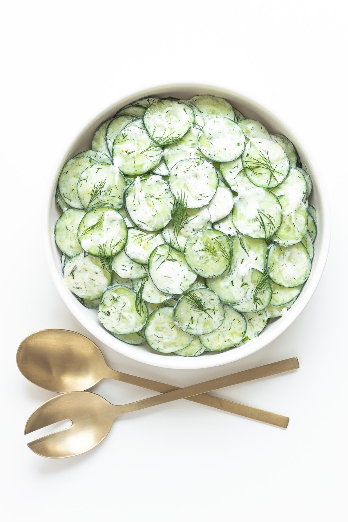 White bowl of cucumber yogurt salad with gold serving spoons.