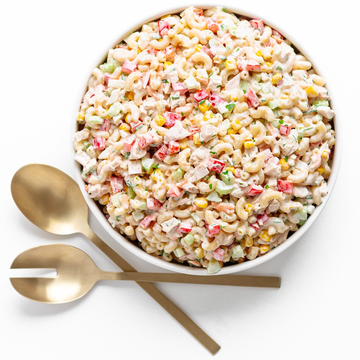 Bowl of macaroni chicken salad next to gold colored salad servers on a white background.