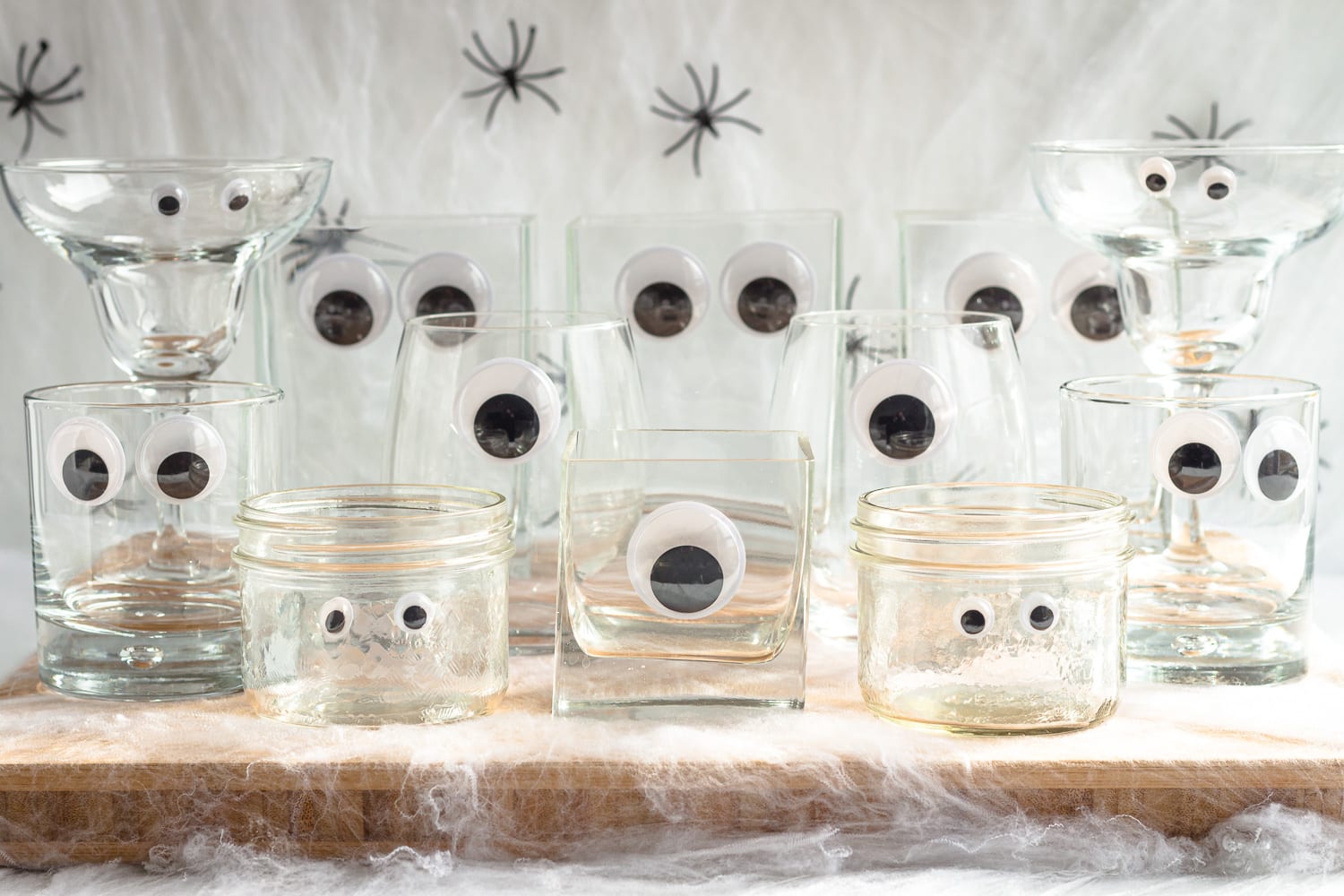 Variety of glassware with craft googly eyes arranged on a wooden board surrounded by fake cobwebs and spiders.