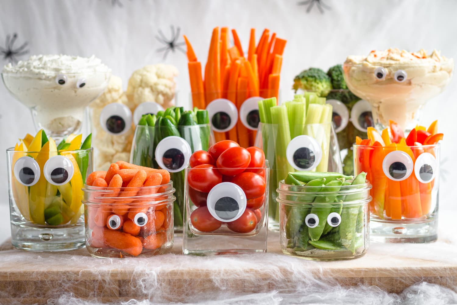A monster Halloween themed veggie tray featuring a variety of glassware with googly eyes filled with different veggies and dip.