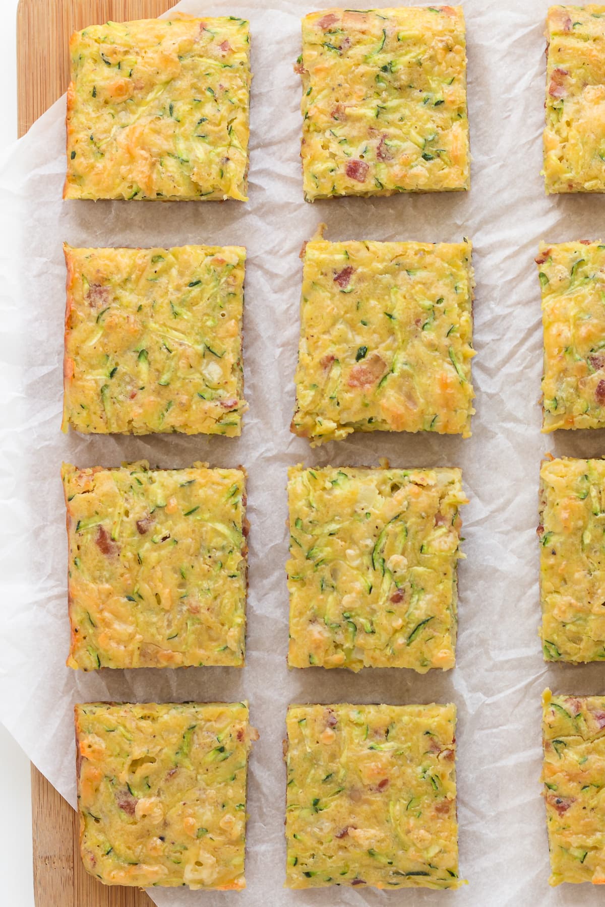 Closeup of square pieces of zucchini slice on a wood board lined with a piece of parchment paper.
