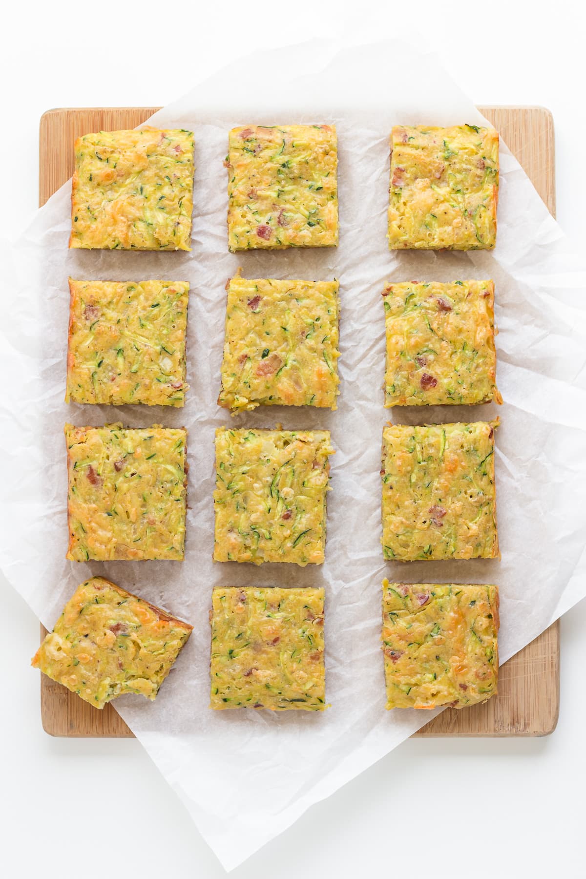 Square cut up pieces of zucchini slice on a wooden board lined with parchment paper.