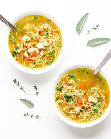 Two bowls of turkey soup on a white background with sage and thyme scattered.