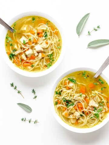 Two bowls of turkey soup on a white background with sage and thyme scattered.