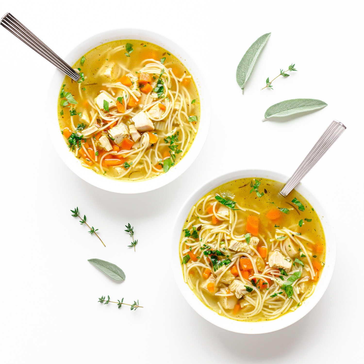 Two bowls of turkey soup on a white background with sage and thyme scattered.