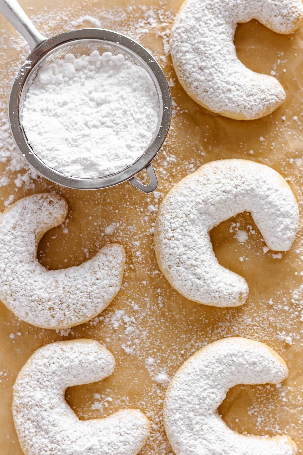 German Vanillekipferl on parchment paper with a small fine mesh sieve filled with powdered sugar.