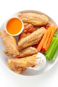 Crispy baked turkey wings on a white plate with carrot and celery sticks and smalls bowls of buffalo wing sauce and blue cheese dip.