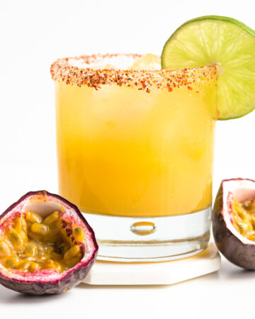 A glass of passion fruit margarita garnished with a lime slice and two passion fruit halves sitting beside it.