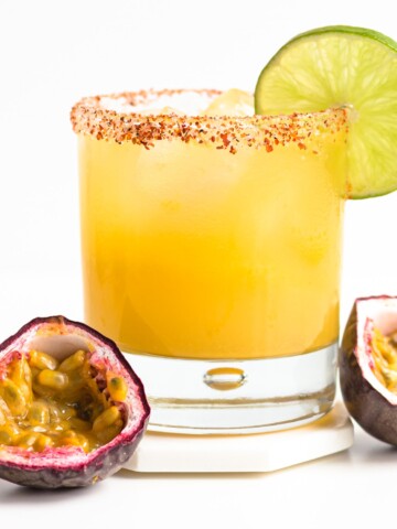 A glass of passion fruit margarita garnished with a lime slice and two passion fruit halves sitting beside it.