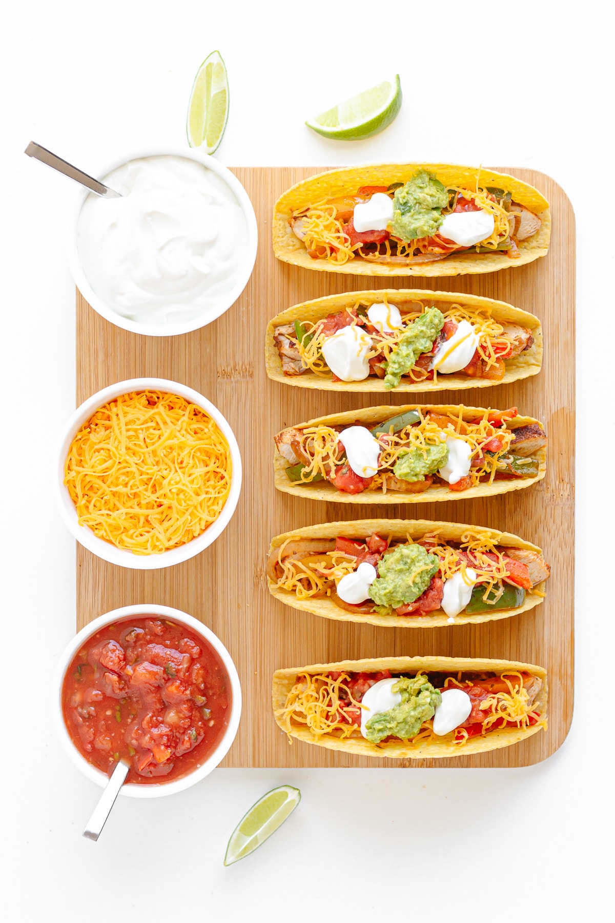 Wooden board with five crunchy fajita tacos and small bowls of sour cream, shredded cheese and salsa on top.