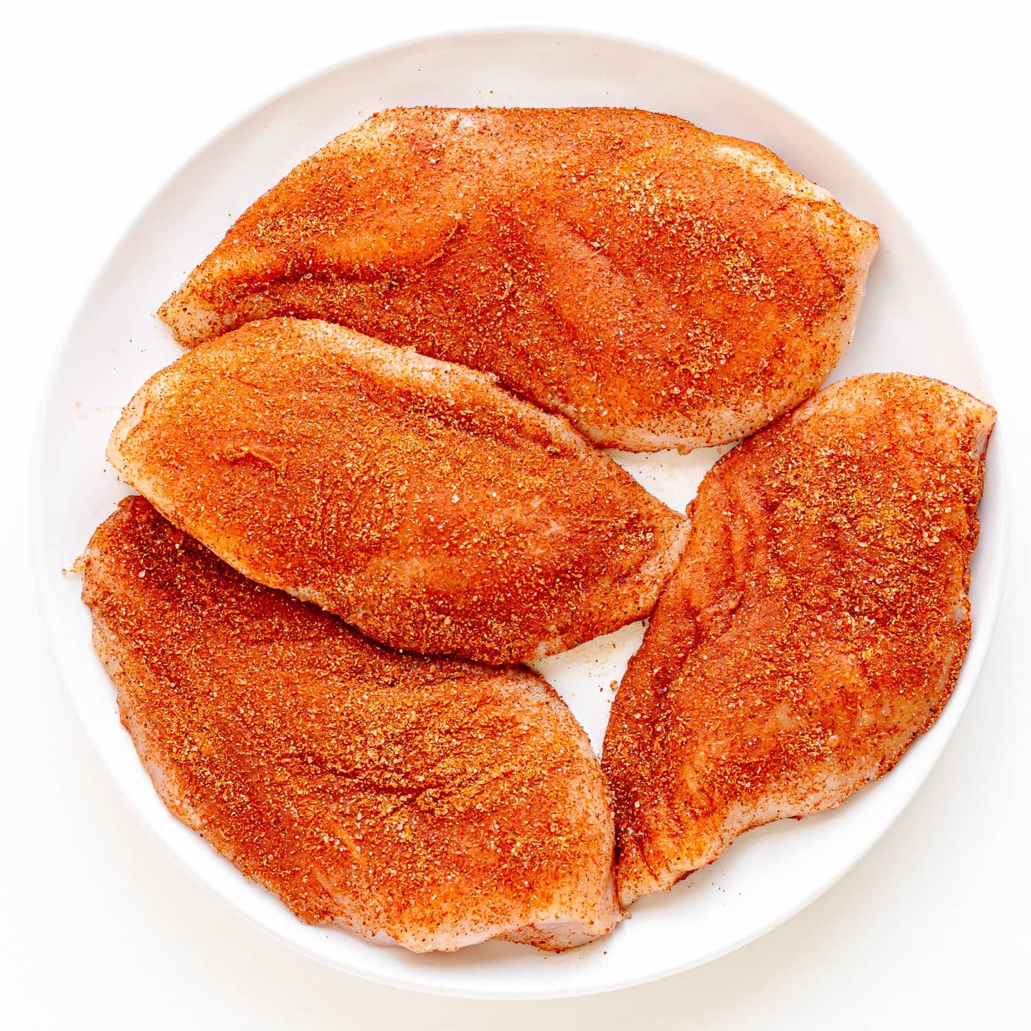Four raw chicken breasts that have been seasoned with fajita seasoning on a white plate.