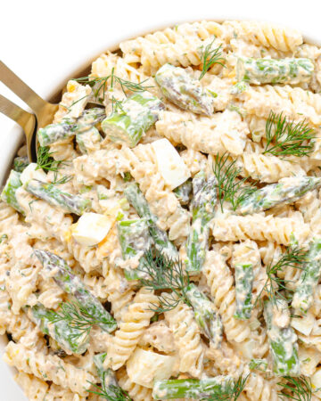 Salmon and asparagus pasta salad with a creamy lemon dill dressing in a white serving bowl with gold salad servers.