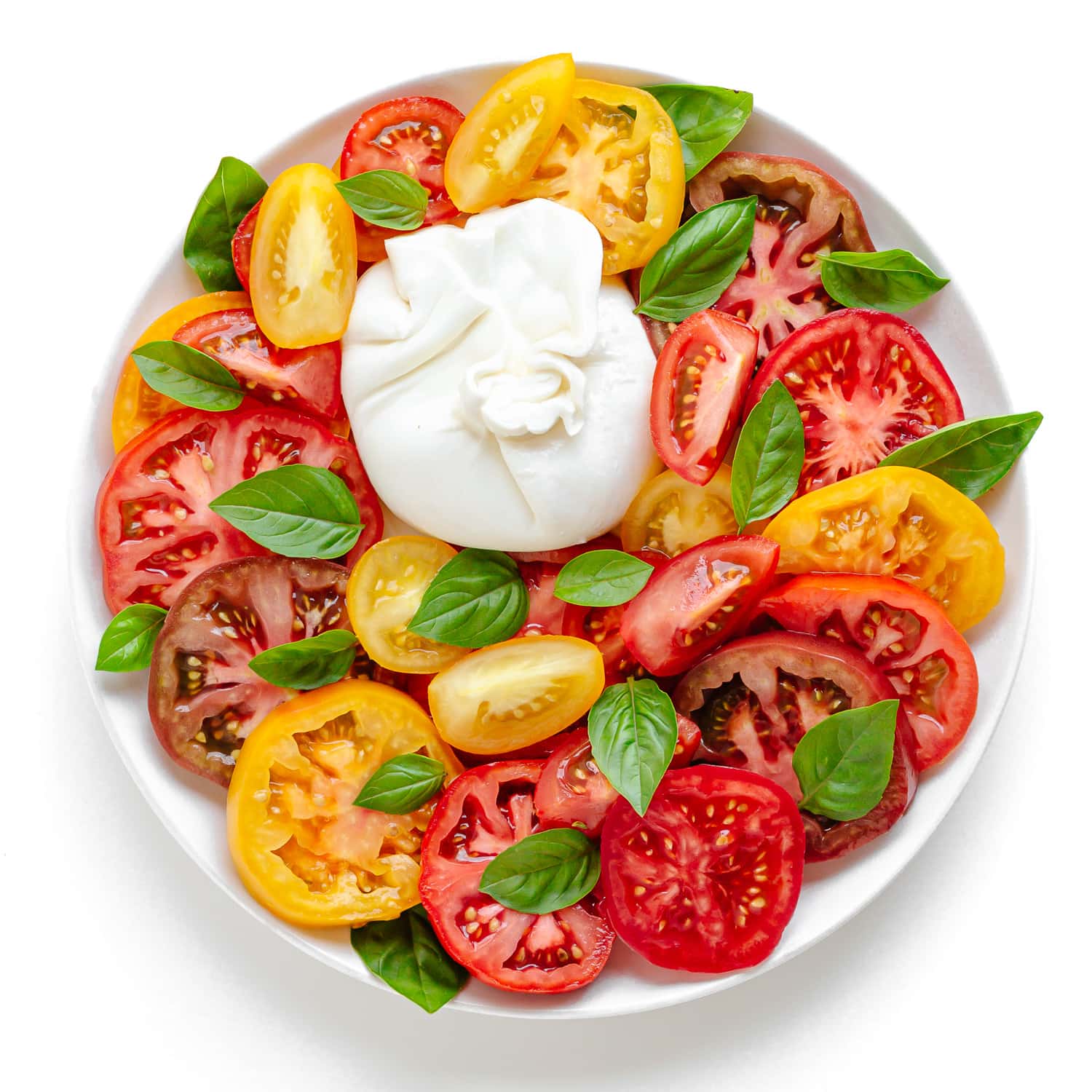 Burrata, sliced heirloom tomatoes and basil assembled on a white plate.