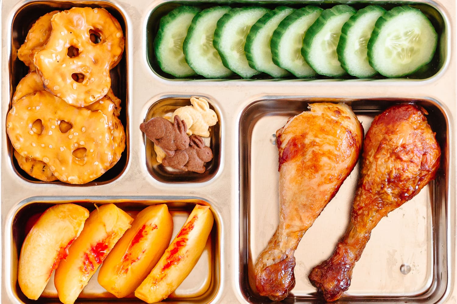 Kids bento lunch box with two roasted chicken drumsticks, cucumber slices, peach slices, pretzel crackers and small bunny shaped cookies.