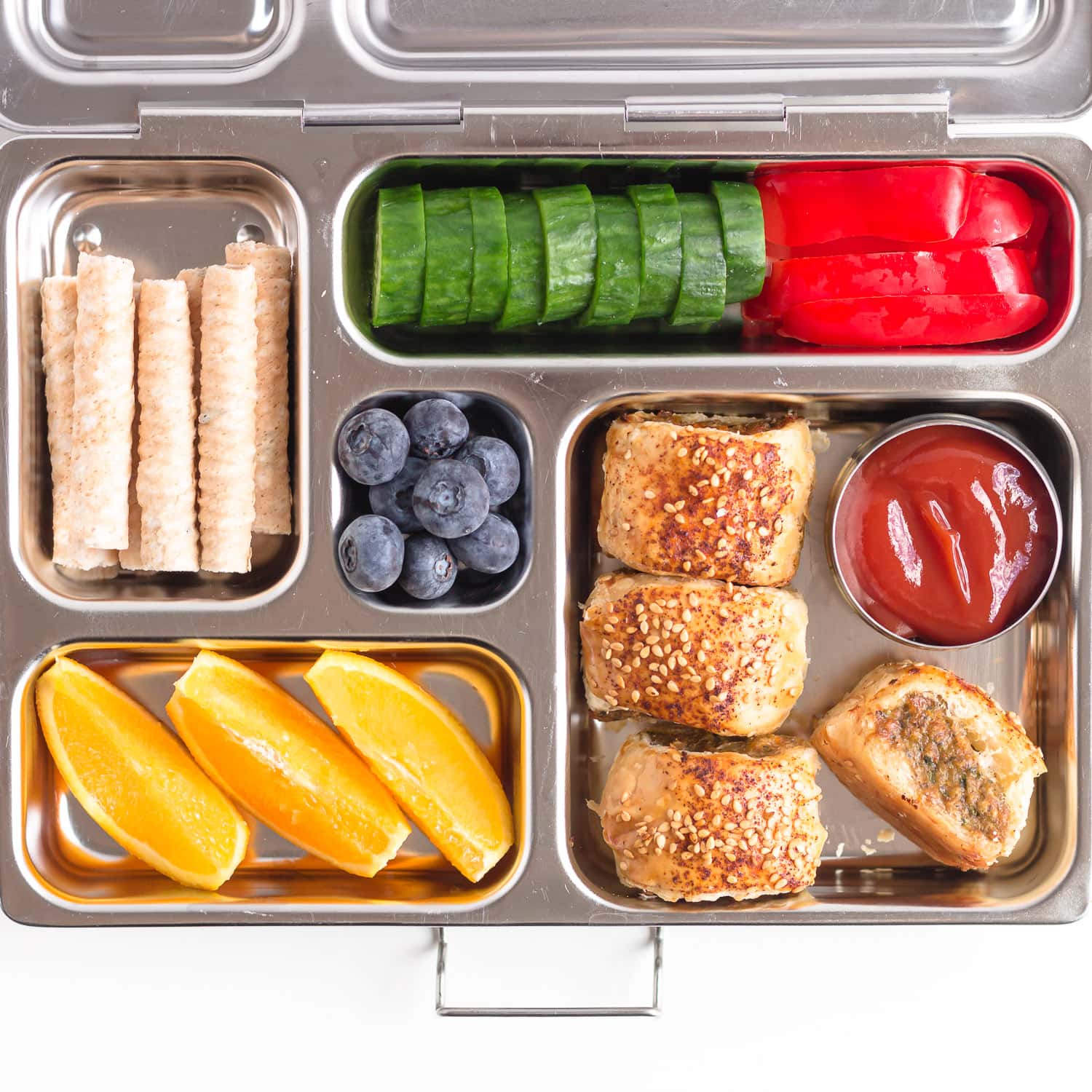 Bento style stainless steel lunch box filled with sausage rolls, orange slices, blueberries, cucumber, red pepper and coconut cookies.