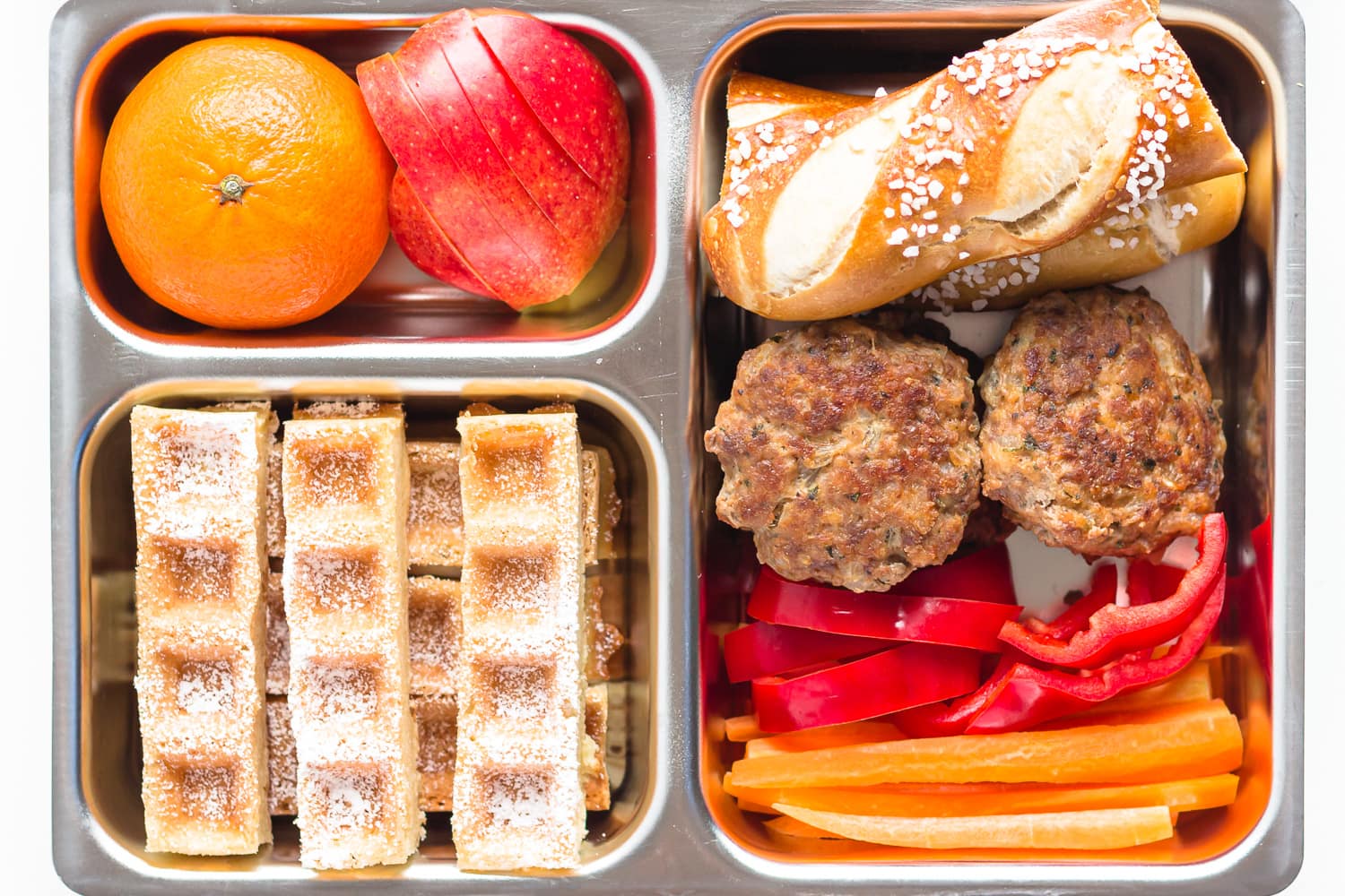 Lunch box filled with cold frikadellen, pretzel stick, red bell pepper slices, carrot sticks, sliced german waffles, apple and mandarin.
