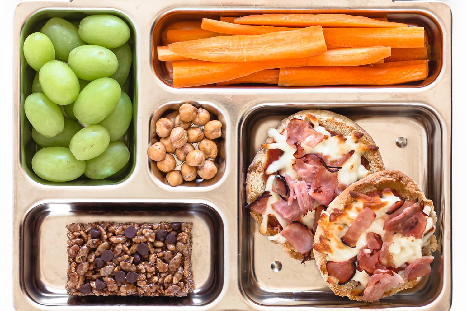 Kids lunch box filled with english muffin pizzas, carrot sticks, green grapes, roasted chickpeas and snack bar.