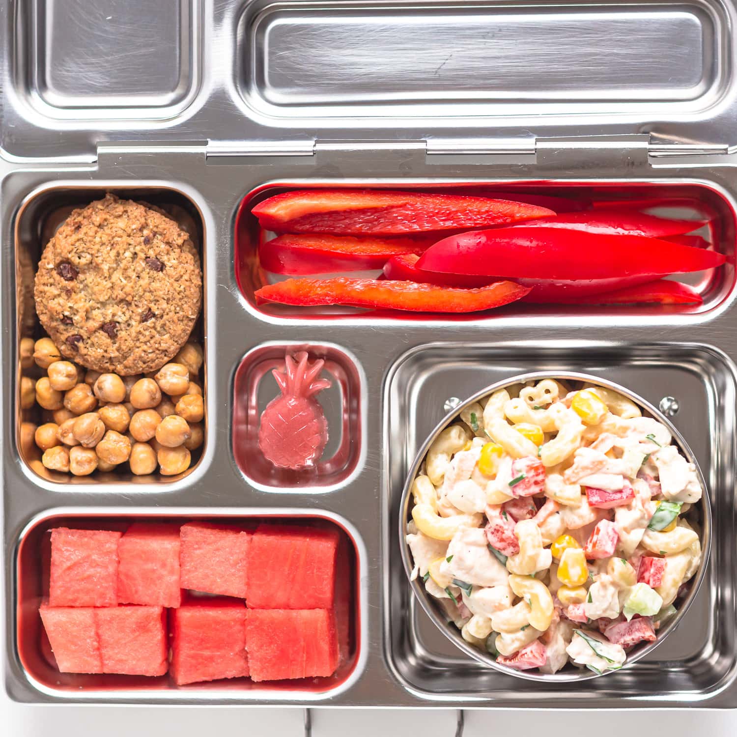 Lunch box containing chicken macaroni pasta salad, cubed watermelon, sliced red bell pepper, strawberry gummy, cookie and roasted chickpeas.