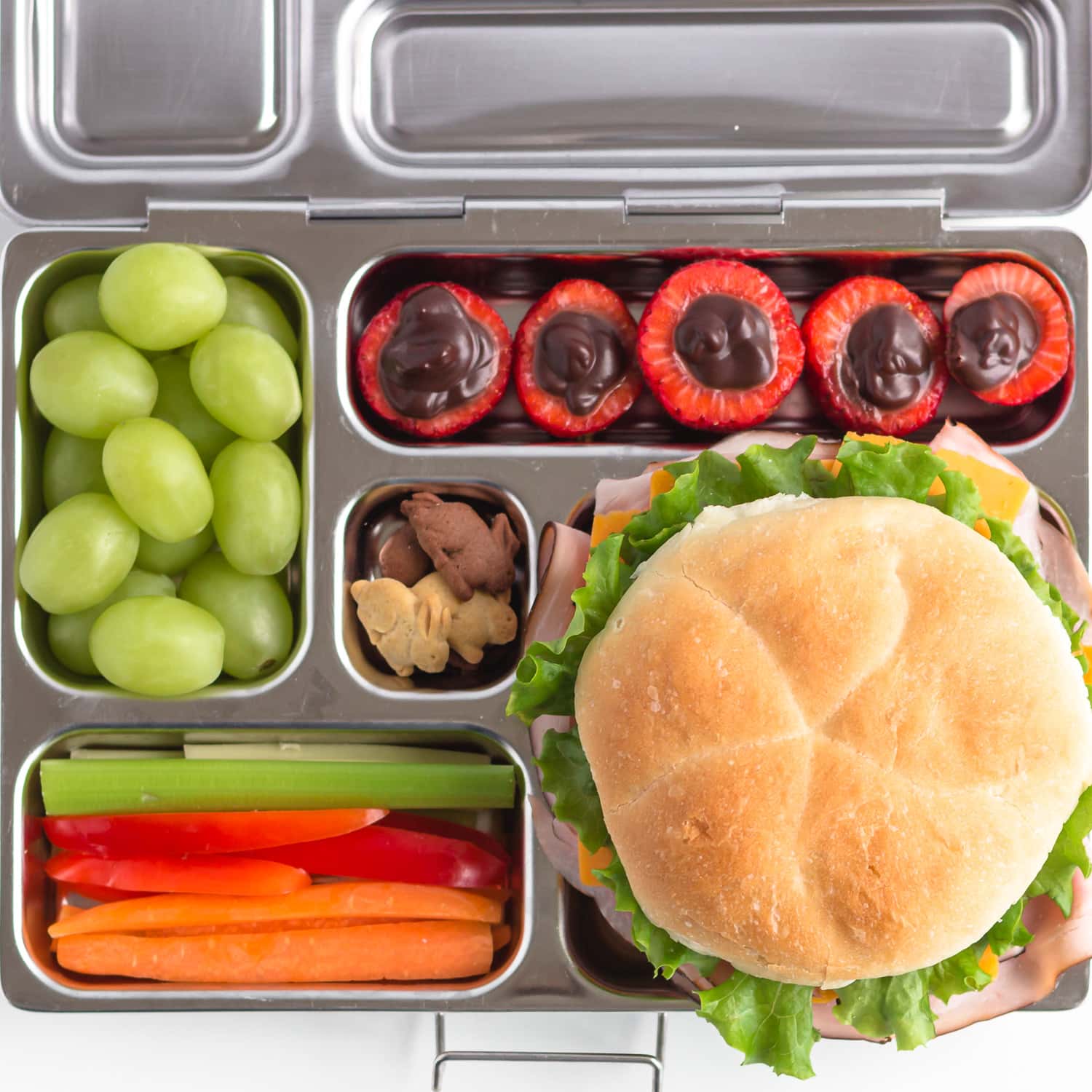 Stainless steel bento-style lunch box filled with a bun sandwich, grapes, veggie sticks, chocolate filled strawberries and small bunny cookies.