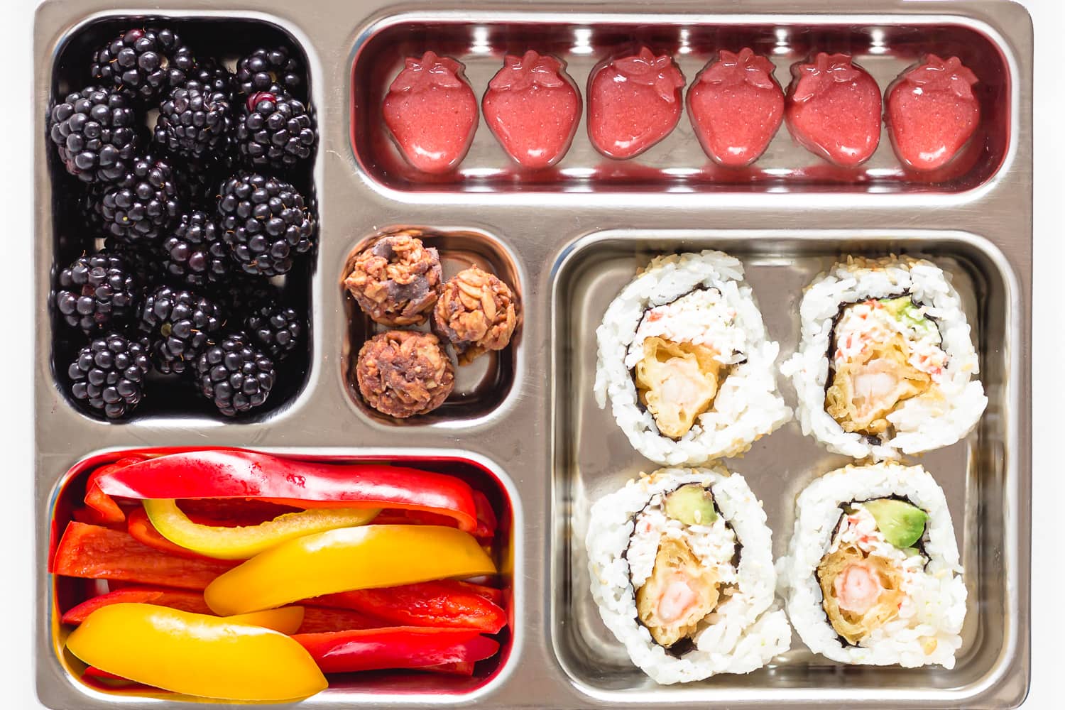 Bento lunch box filled with sushi rolls, bell pepper slices, blackberries, strawberry gummies and granola bites.