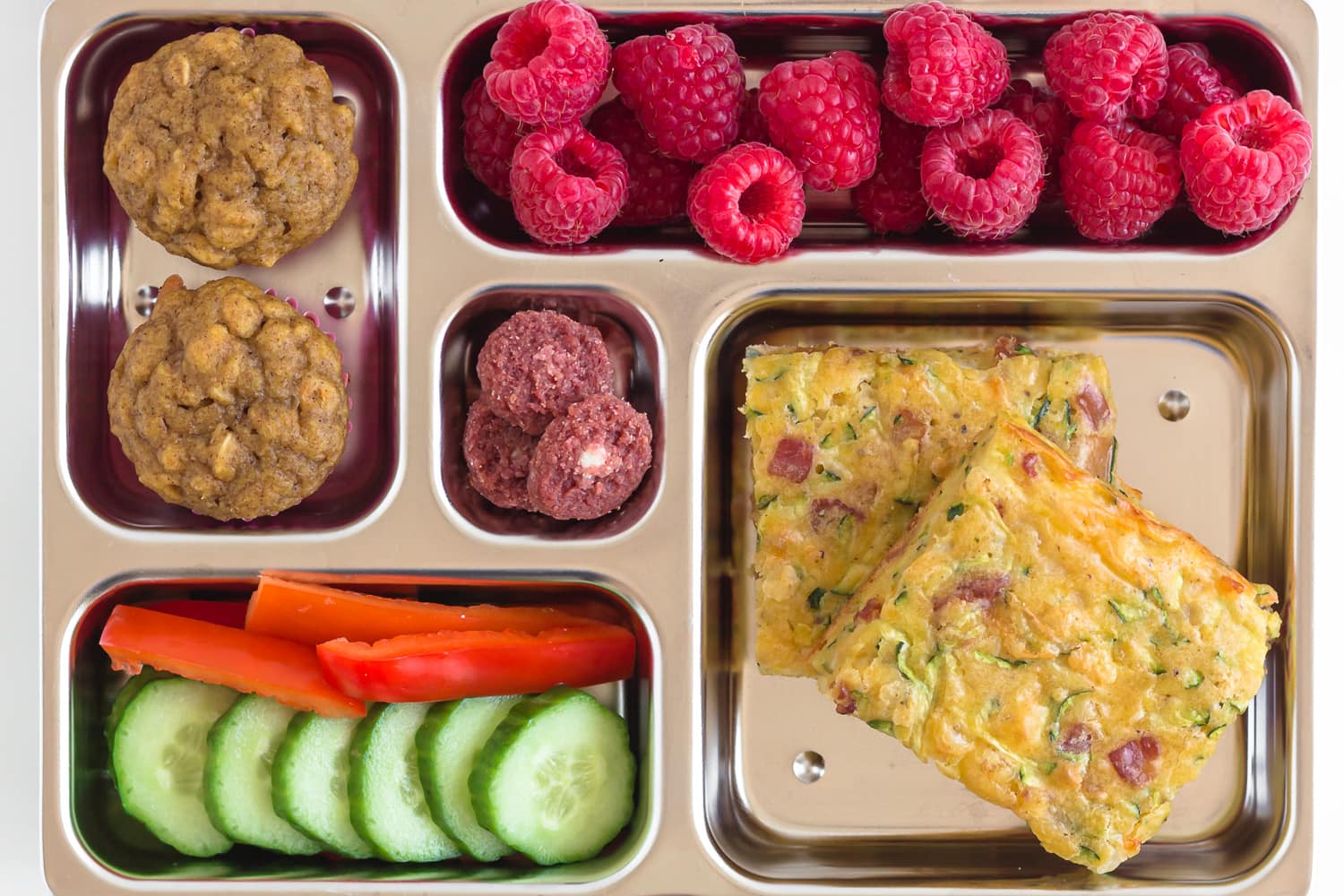 Stainless steel lunch bento box containing zucchini slice, raspberries, cucumber and red bell pepper slices, mini muffins and mini cookies.