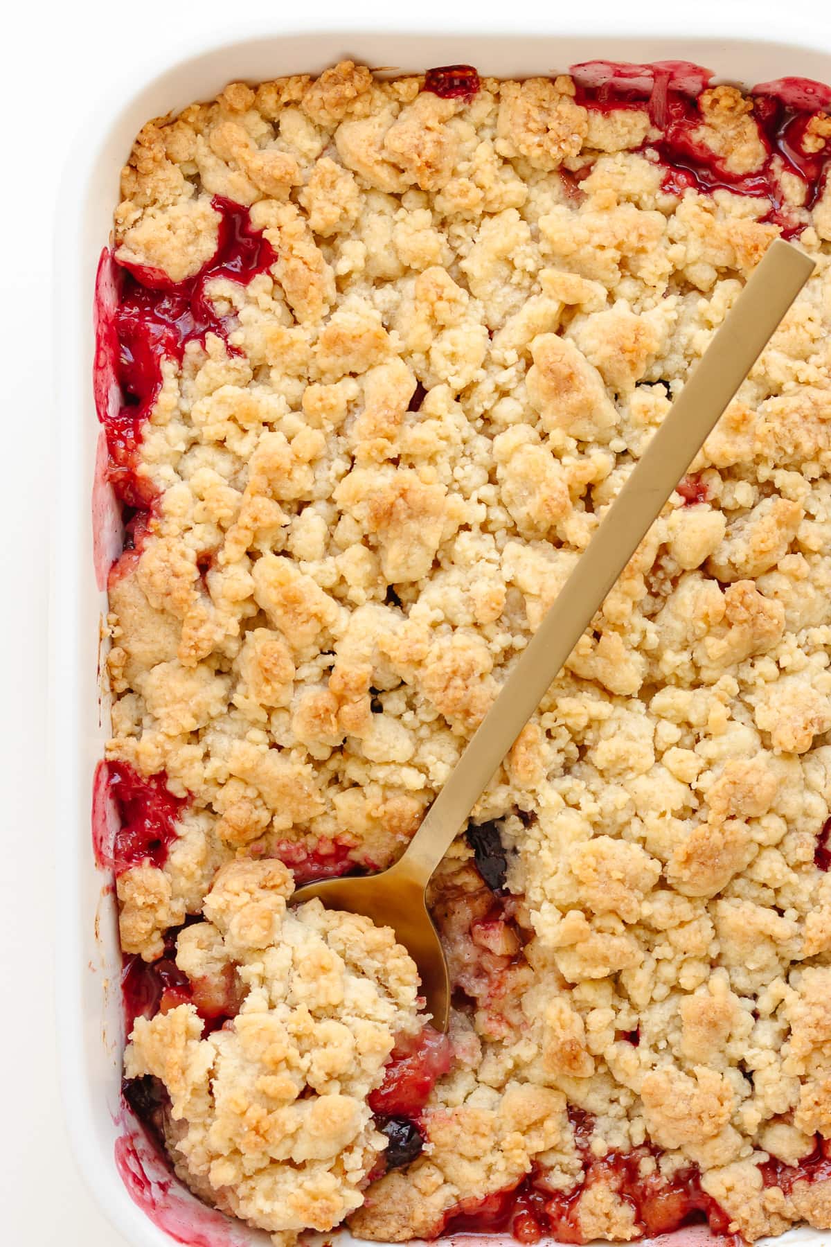 Apple and plum crumble in a white baking dish with gold colored serving spoon.