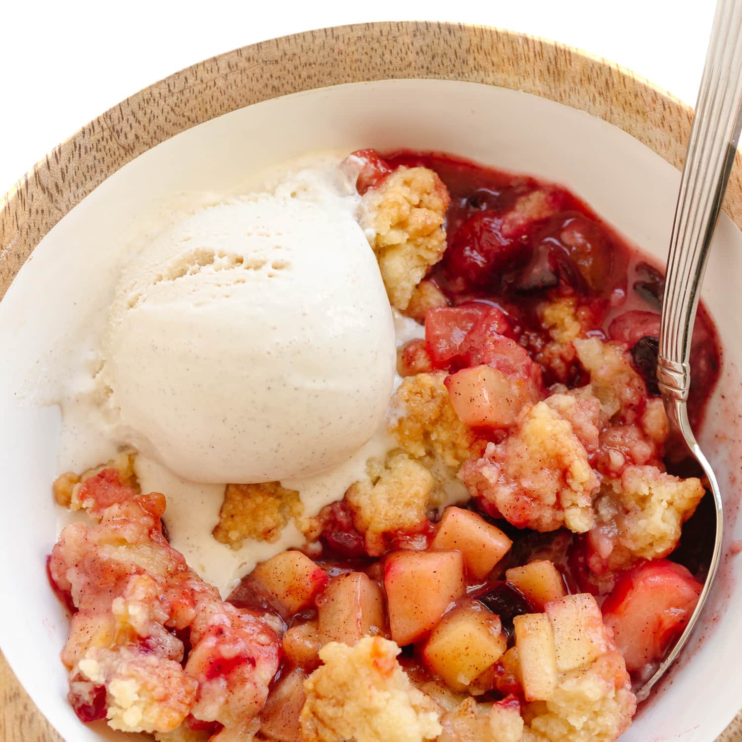 Bowl of plum and apple crumble with a spoon and scoop of vanilla ice cream melting on top.