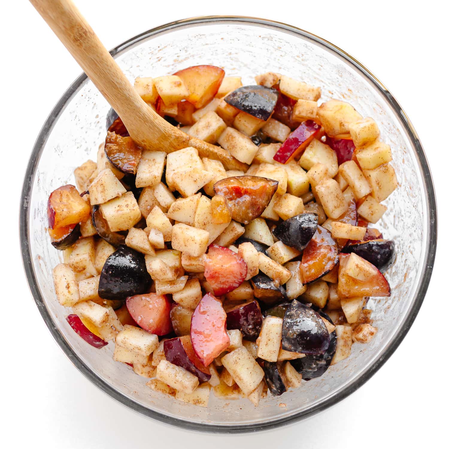 Plum and apple fruit mixture for crumble being mixed in a glass bowl with a wooden spoon.