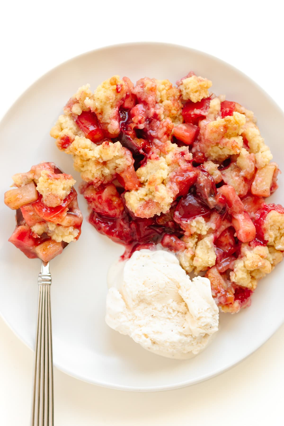 Plum and apple crumble on a white plate with a scoop of vanilla ice cream and spoon.