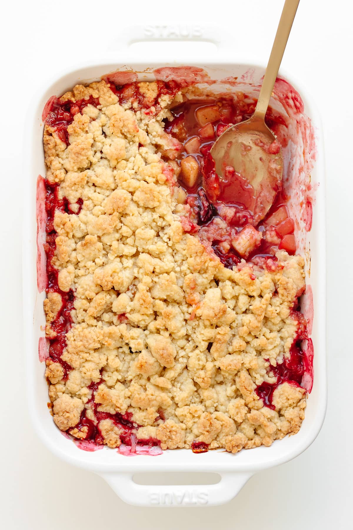 Plum apple crumble in a white baking dish with a serving spoon sitting in a corner where a portion has been scooped out.