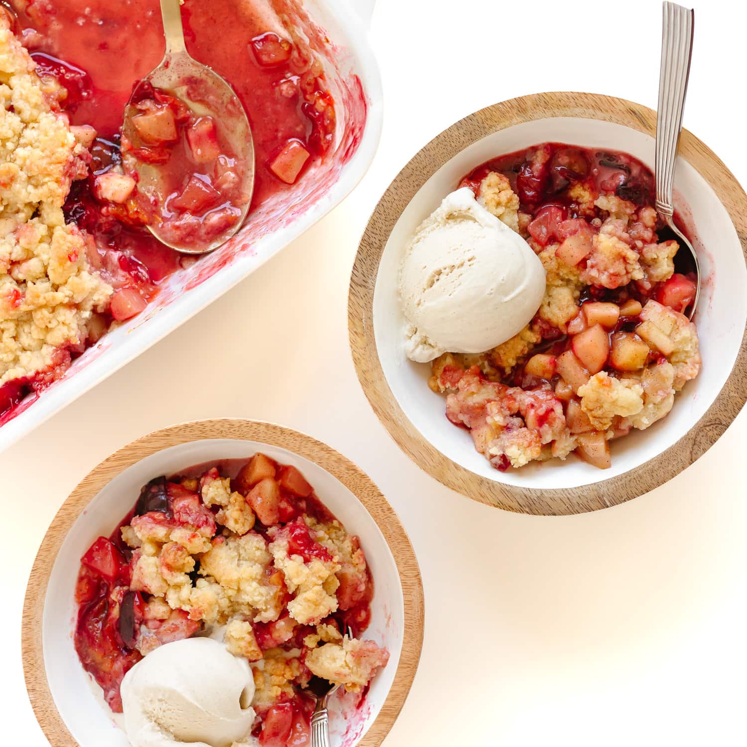 Plum apple crumble being served into two bowls with a scoop of vanilla ice cream.