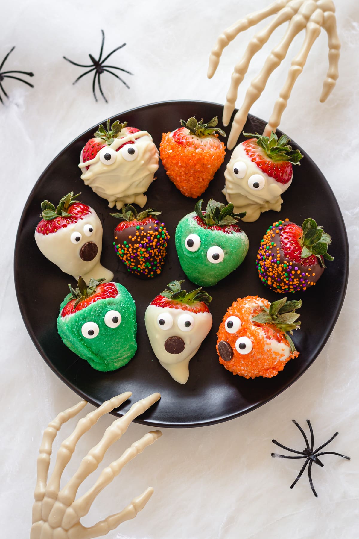 A variety of Halloween chocolate covered strawberries on a black plate with two skeleton hands reaching in.