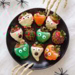 A variety of different Halloween strawberries on a black plate with two skeleton hands reaching in.