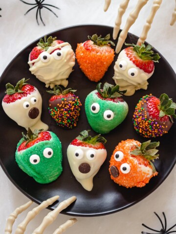 A variety of different Halloween strawberries on a black plate with two skeleton hands reaching in.