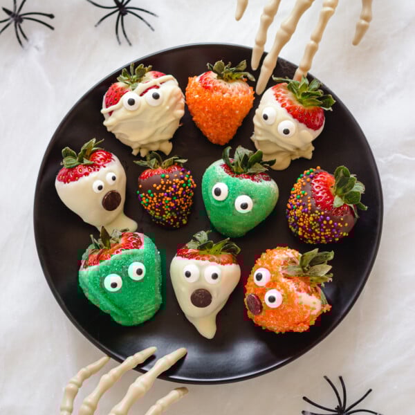 A variety of different Halloween strawberries on a black plate with two skeleton hands reaching in.