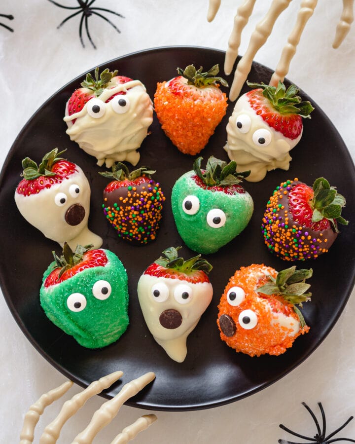 A variety of different Halloween strawberries on a black plate with two skeleton hands reaching in.