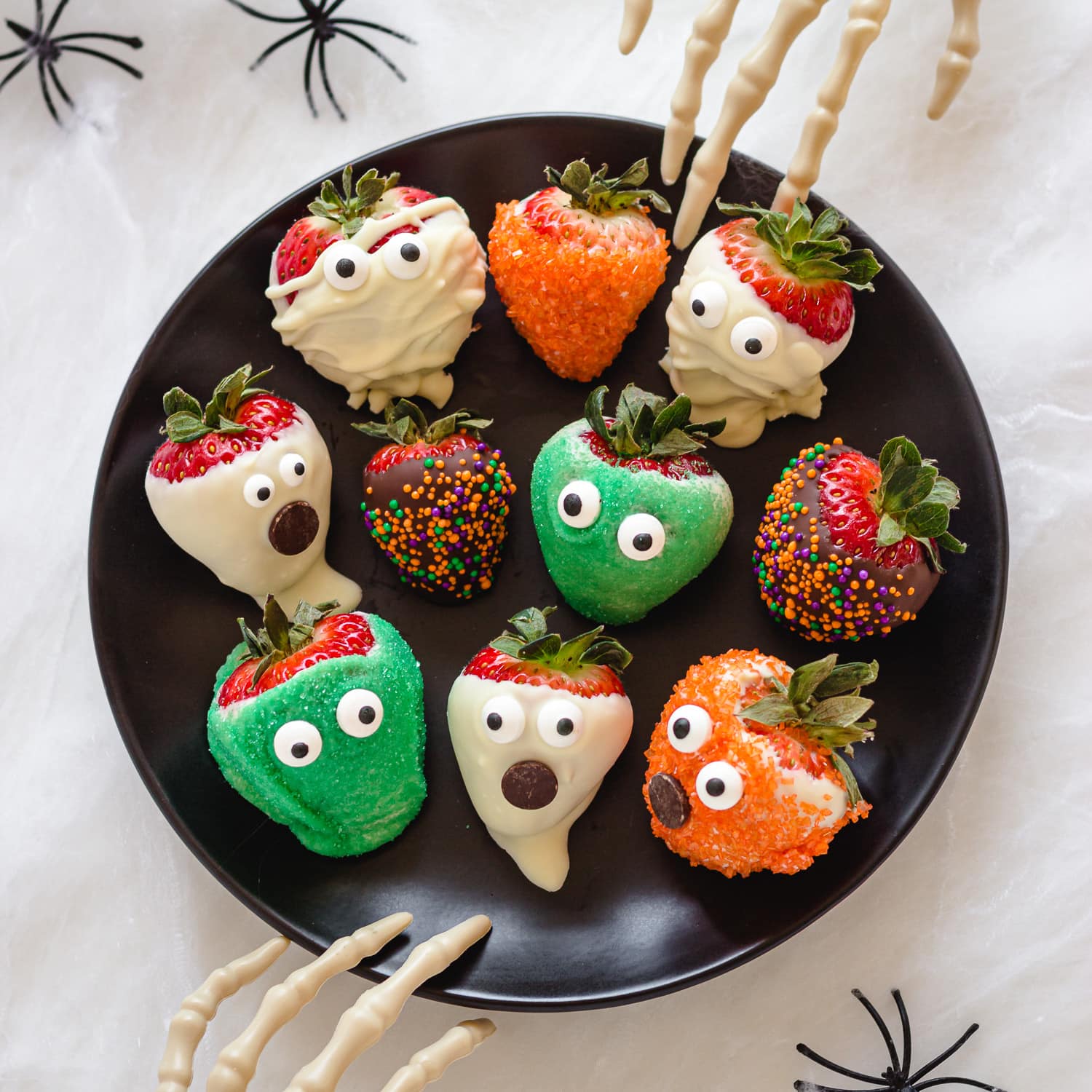 A variety of different Halloween strawberries on a black plate with two skeleton hands reaching in.