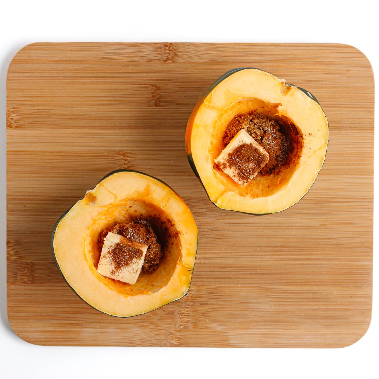 Butter and brown sugar topping distributed between two acorn squash halves on a wooden cutting board.