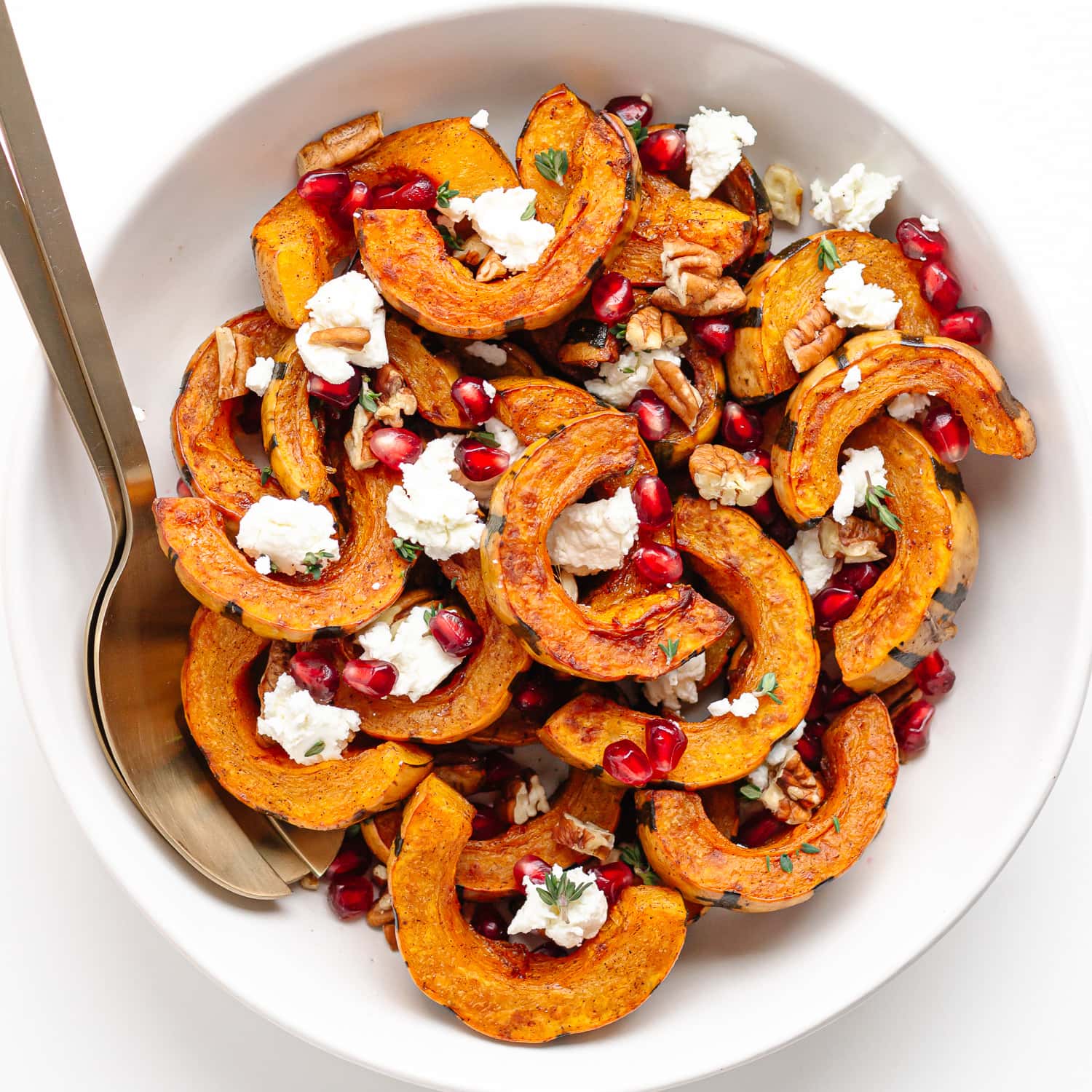 Roasted delicata squash slices in a white serving bowl garnished with pomegranate arils, pecans, thyme and goat cheese.