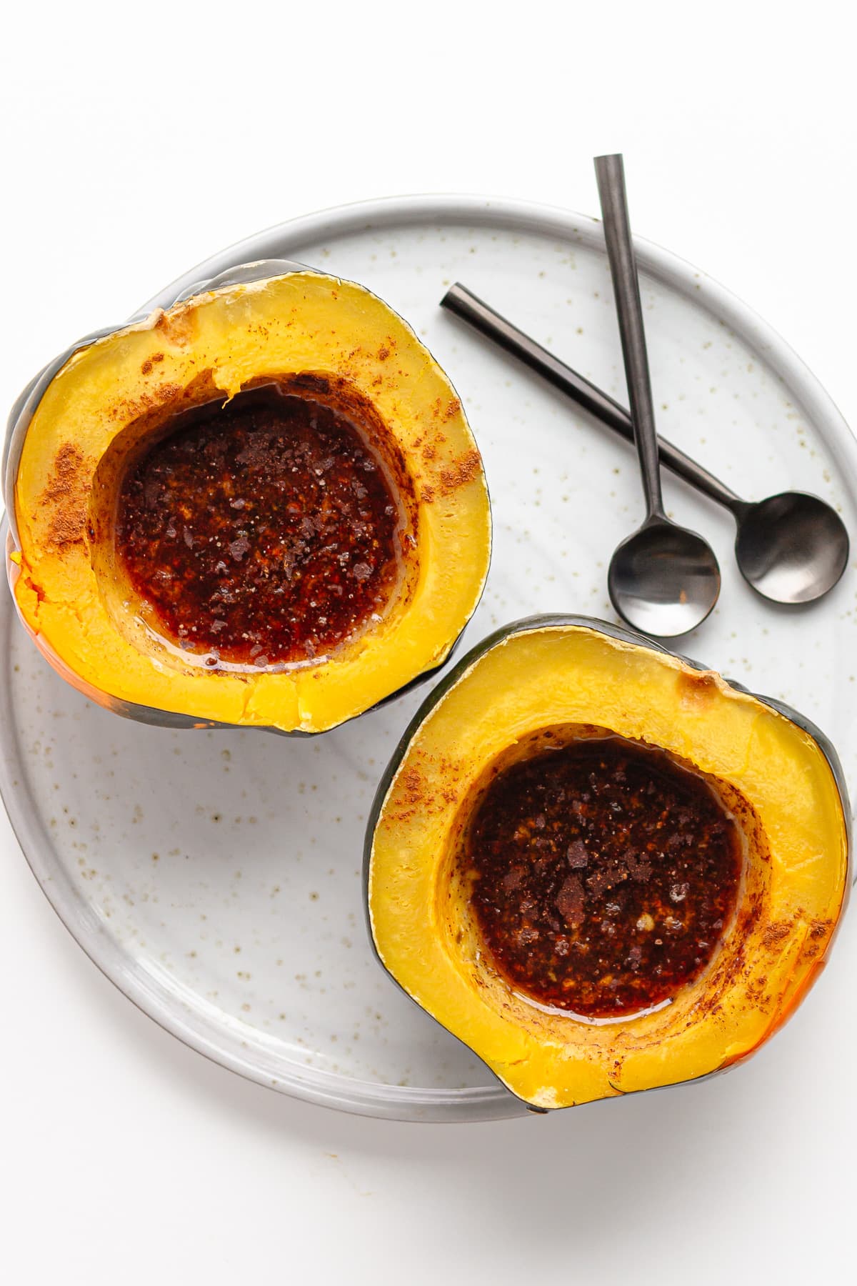 Two acorn squash halves filled with butter, brown sugar and cinnamon on a serving plate with two spoons.