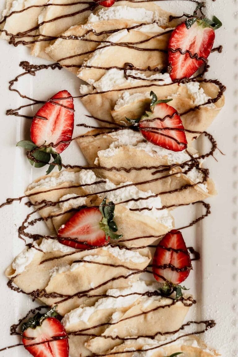 Cream filled crepes on a white platter that are drizzled with chocolate and garnished with strawberries.