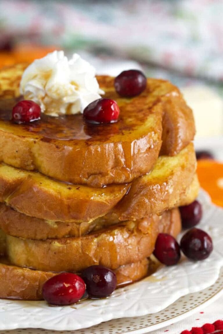 Stack of eggnog french toast on a white plate topped with cranberries, maple syrup and whipped cream.