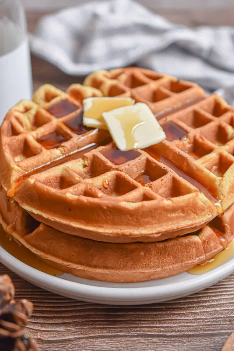 Two eggnog waffles stacked on a white plate and topped with maple syrup and two pats of butter.