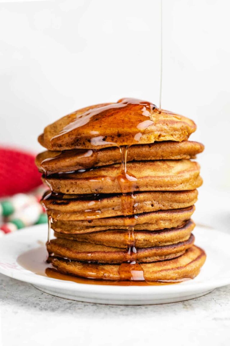 Stack of gingerbread pancakes on a white plate with syrup being poured over top.